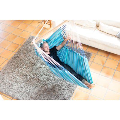 A person reclining in a blue striped hammock suspended over a gray shag rug on a tiled floor, with a white sofa in the background.