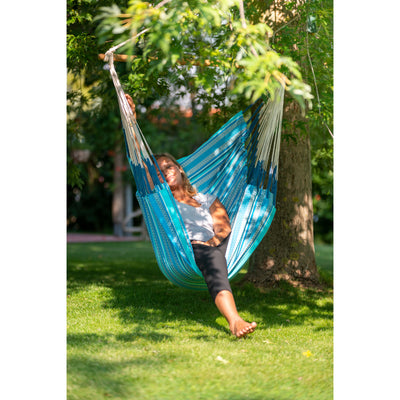 A woman relaxes in a blue and white striped woven hammock suspended between two trees on a sunny green lawn.
