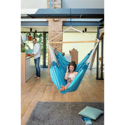 A woman relaxes in a blue and white striped fabric hammock suspended from a wooden bar in a modern open-plan loft with wood floors and brick columns. A man is visible in the background near a kitchen counter.