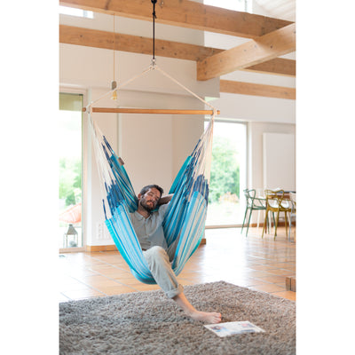 A man relaxes in a hanging fabric hammock suspended from a wooden bar in a bright, modern room with exposed wood ceiling beams and tiled floors.