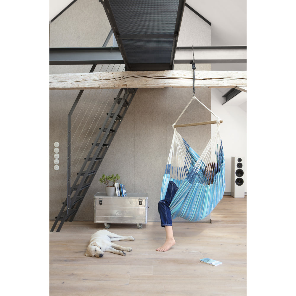 A person relaxes in a blue and white striped hanging hammock suspended from a wooden loft staircase in a modern loft apartment. A white dog is lying on the wooden floor nearby, and a small silver storage chest sits against the wall.