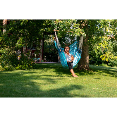 A person sitting in a bright blue and white striped hammock suspended between two trees on a grassy lawn, with green foliage and a house visible in the background.
